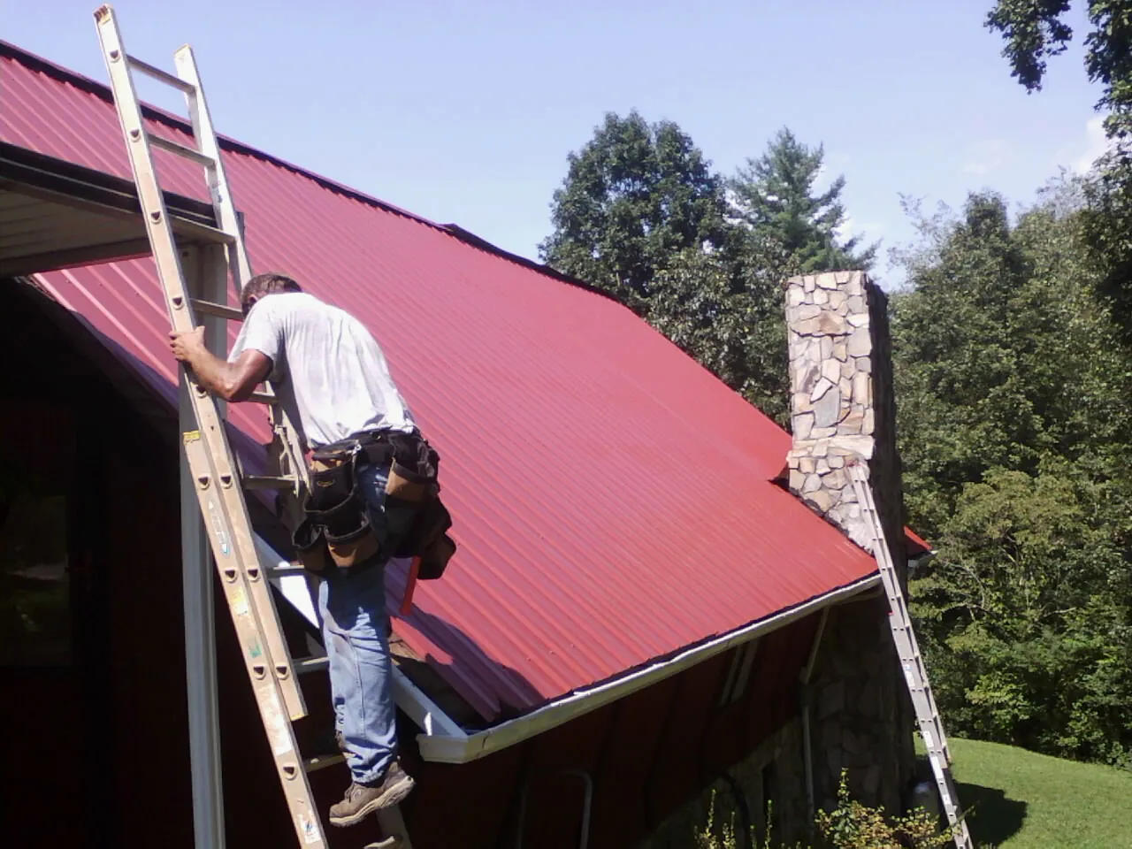 Roofer on a ladder beside a red metal roof and stone chimney — Soffit & Fascia Repair by Roofing Brothers in Farnham